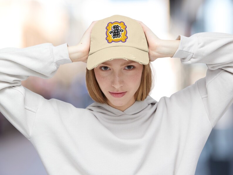 K&ouml;nnte beinhalten: Eine beige Baseballkappe mit abgenutztem Look. Die Vorderseite der Kappe zeigt einen gestickten Aufn&auml;her in Gelb und Lila mit dem Text "I'll be there for you" in einem lila Rahmen.