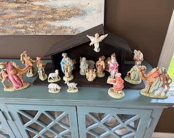 May include: A Christmas Nativity scene with figurines of Mary, Joseph, the Three Wise Men, shepherds, angels, and animals. The figurines are on a blue cabinet, with a brown wooden stable in the background.