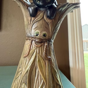 May include: A whimsical, vintage-style ceramic lamp featuring a black cat perched atop a stylized wheat stalk. The lamp has a detailed, textured design with green and brown hues, and cartoonish eyes. The lamp is a unique decorative item.