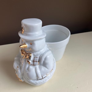 May include: White ceramic snowman planter with gold accents. The snowman is wearing a hat and has a bow tie. The planter has a removable top.