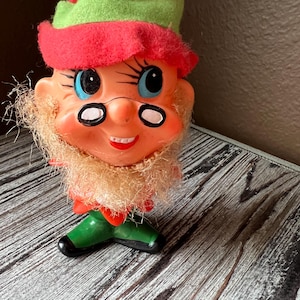 May include: A vintage Christmas ornament shaped like an elf's head. The elf has a red hat with a green brim, a white beard, and a big smile. The ornament is made of plastic and has a shiny finish.