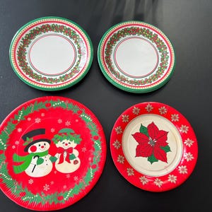 May include: Four paper plates with a Christmas theme. Two plates feature a green and white border with red holly and berries. One plate has a red background with a snowman and a girl in a green dress. The last plate has a red poinsettia on a white background.