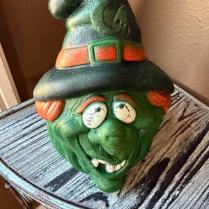 May include: A green Halloween witch head with a black hat, orange belt, and white eyes. The witch has a wide grin and orange hair. The witch is a decorative item, perfect for Halloween.
