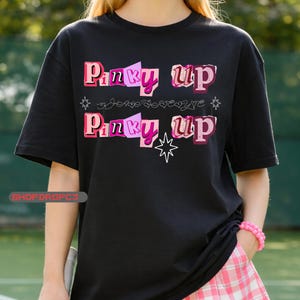 May include: Black t-shirt with the text "Pinky Up" in pink and purple block letters. The shirt also features a star and other decorative elements. The model is wearing a pink bracelet and a pink and white plaid skirt.
