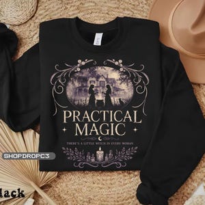 May include: A black long-sleeve sweatshirt featuring a graphic print of a house, two figures, and a cat. The text reads PRACTICAL MAGIC and THERE'S A LITTLE WITCH IN EVERY WOMAN, accompanied by illustrations of a candle, bottles, and floral flourishes.
