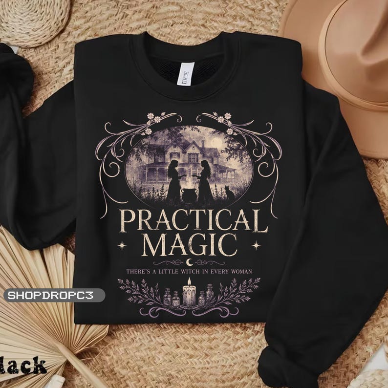 May include: A black long-sleeve sweatshirt featuring a graphic print of a house, two figures, and a cat. The text reads PRACTICAL MAGIC and THERE'S A LITTLE WITCH IN EVERY WOMAN, accompanied by illustrations of a candle, bottles, and floral flourishes.