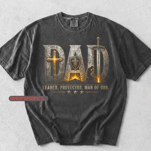 May include: Dark gray t-shirt with the word "DAD" in a distressed, textured design. The design includes a cross, shield, sword, and the text "LEADER. PROTECTOR. MAN OF GOD." The shirt has a vintage, worn look.