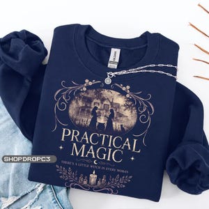 May include: A navy blue crewneck sweatshirt featuring a graphic print of a house, two figures, and a cat. The text reads "PRACTICAL MAGIC" and "THERE'S A LITTLE WITCH IN EVERY WOMAN" with decorative floral borders and a candle illustration.
