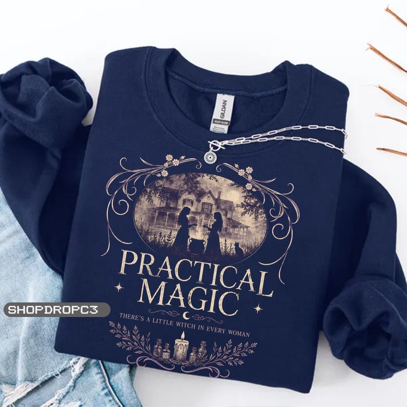 May include: A navy blue crewneck sweatshirt featuring a graphic print of a house, two figures, and a cat. The text reads "PRACTICAL MAGIC" and "THERE'S A LITTLE WITCH IN EVERY WOMAN" with decorative floral borders and a candle illustration.