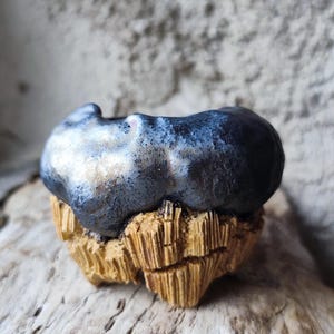 Peut inclure: Un petit bol en céramique présentant une partie supérieure lisse, bleu métallique et gris anthracite, avec un bord irrégulier et organique. La base est texturée avec un aspect bois beige, marqué par des stries verticales et des fissures.