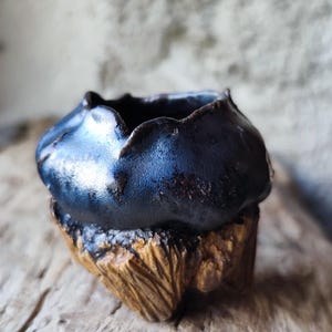 Könnte beinhalten: Eine kleine, dunkelblaue Keramikschale mit strukturierter Oberfläche, die auf einem geschnitzten Holzsockel ruht. Der Rand der Schale hat eine einzigartige, wellenförmige Form. Der Holzsockel hat ein natürliches, rustikales Aussehen.