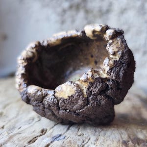 Op de afbeelding: Een kleine, handgemaakte keramische kom met een ruwe, getextureerde buitenkant. De kom is voornamelijk donkerbruin met lichtere bruin- en crèmekleurige accenten rond de rand. Hij staat op een houten oppervlak, wat de organische vorm benadrukt.
