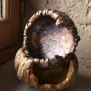 Peut inclure: Un bol en céramique fait à la main, en deux parties, avec un design texturé et organique. Le bol supérieur a un intérieur sombre et irisé, tandis que la base et l'extérieur présentent une texture rugueuse et terreuse.