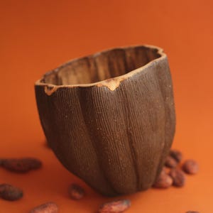 May include: A dark brown ceramic cup with a textured surface, sitting on a brown background with scattered cocoa beans.