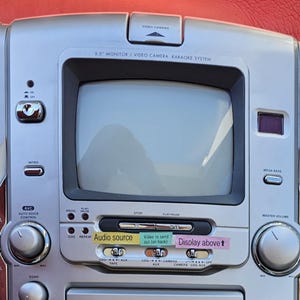 Könnte beinhalten: Silberne Memorex Karaoke-Maschine mit eingebautem 14 cm Monitor. Merkmale sind ein Kassetten-Player/Recorder, Mikrofoneingänge und verschiedene Bedienknöpfe. Der Text "KARAOKE RECORDING" ist sichtbar.