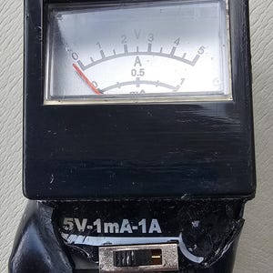 May include: A black analog voltmeter with a clear display showing voltage and amperage readings. The meter has a needle and a scale from 0 to 5, with text indicating 5V-1mA-1A. A switch is visible below the display.