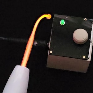 Puede incluir: Primer plano de un dispositivo de tratamiento de la piel de alta frecuencia. El dispositivo tiene un mango blanco y un electrodo de vidrio curvo y naranja brillante. Una caja de control negra con una luz verde y un dial está conectada al dispositivo.