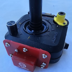 May include: A black joystick with a red and black base. The joystick has a yellow button on the top and a black knob on the side. The base has the text "U2 ALARMIC" on it.
