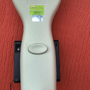 May include: A handheld barcode scanner with a light gray body and a black top section. The device features a central oval button, a Made in China label, a barcode with the text BG7076962, and a metallic connector at the base with a red and black wire.