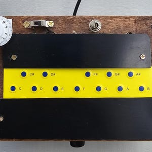 Könnte beinhalten: Ein rechteckiges elektronisches Musikinterface aus Holz mit einer zentralen gelben Platte, die blaue runde Tasten mit den Notenbezeichnungen C, C#, D, D#, E, F, F#, G, G#, A, A# und B aufweist. Es enthält ein weißes nummeriertes Einstellrad, einen Kippschalter und eine rote Kontrollleuchte.