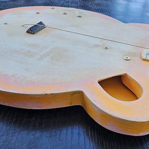 Pode incluir: Uma guitarra elétrica artesanal, inacabada, com corpo em madeira clara e acabamento pintado em laranja e amarelo. A guitarra apresenta um botão de tom branco, uma ponte preta e uma escala com marcações coloridas.