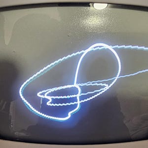 May include: A glowing white and teal light trail forming a continuous, complex looped pattern on a dark, curved screen. The luminous line creates an abstract, overlapping geometric shape with a distinct, jagged texture along the edges of the path.