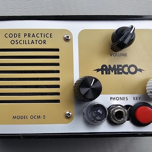May include: Vintage Ameco Model OCM-2 Code Practice Oscillator. The device has a black frame with a cream-colored faceplate. It features a speaker, multiple knobs, and the text "CODE PRACTICE OSCILLATOR" and "AMECO".