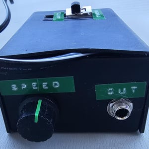 Op de afbeelding: Zwart elektronisch apparaat met een snelheidsregelkno, gemarkeerd met "SPEED", en een uitgangsbus gemarkeerd met "OUT". Een tuimelschakelaar is zichtbaar aan de bovenkant, met de labels "AUTO" en "CONTROL". Een zwart netsnoer is aangesloten.
