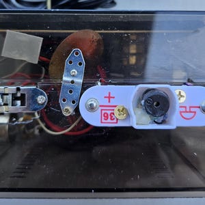 May include: A rectangular, transparent dark plastic electronic housing unit featuring an internal metal slide switch, a small perforated metal bracket, and a white circuit component labeled with a plus sign and the number 36 in red ink.