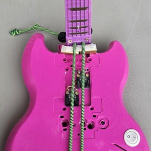 Puede incluir: Una guitarra de juguete desmontada en rosa brillante y morado. El cuerpo es rosa con un interruptor de encendido/apagado blanco y cuerdas verdes y blancas. El mástil es morado con marcadores de traste marrones. A la guitarra le faltan algunos componentes.