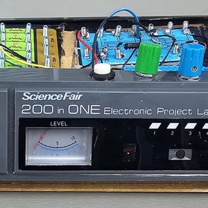 Könnte beinhalten: Ein graues Science Fair 200-in-ONE Electronic Project Lab mit einem weißen Tonregler, einem Bedienknopf und einem Pegelmessgerät. Das Projektlabor hat einen roten Knopf, einen Lautsprecher und eine darin sichtbare Leiterplatte. Ein grüner und blauer Fidget Spinner liegt oben auf.