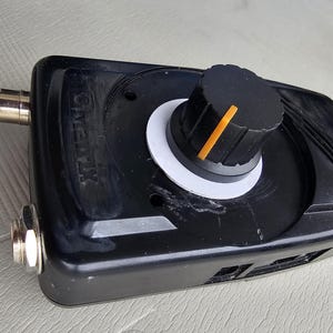 May include: Black electronic device with a large, textured knob featuring an orange indicator line. The device has a white ring around the knob and metallic connectors. The text "VERTEX" is visible on the device.