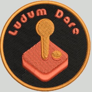 Può includere: Una toppa ricamata nera e arancione con il testo "Ludum Dare" e un'immagine stilizzata di un joystick.