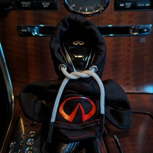 Könnte beinhalten: Schwarzer Automatik-Wählhebel-Knopf mit schwarzer Kapuzenhülle mit rotem Infiniti-Logo auf der Vorderseite. Die Kapuze hat weiße Kordeln.