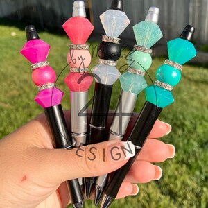 May include: Five colorful pens with clear diamond-shaped tops and silver and black accents. The pens are held in a hand with a white manicure.