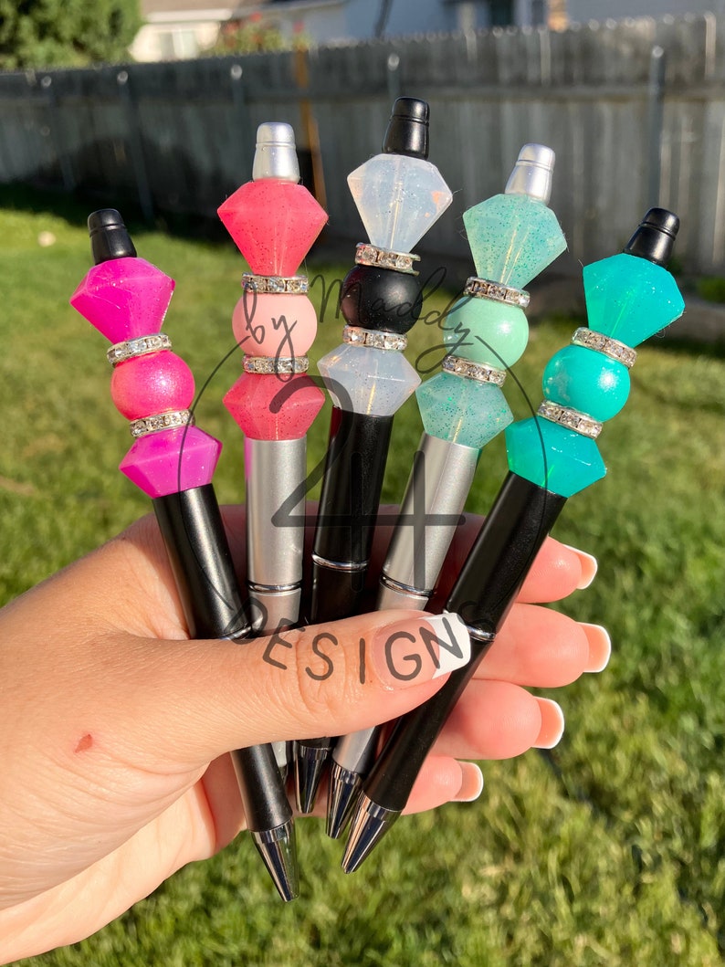 May include: Five colorful pens with clear diamond-shaped tops and silver and black accents. The pens are held in a hand with a white manicure.