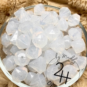 May include: A bowl of white, sparkly, faceted, silicone beads. The beads are shaped like diamonds and have a subtle shimmer. The bowl is on a brown, fuzzy surface. The text "by Maddy 24+ DESIGNS" is visible on the bottom right of the image.