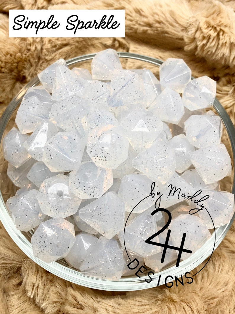 May include: A bowl of white, sparkly, faceted, silicone beads. The beads are shaped like diamonds and have a subtle shimmer. The bowl is on a brown, fuzzy surface. The text "by Maddy 24+ DESIGNS" is visible on the bottom right of the image.