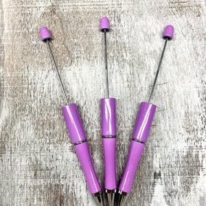 May include: Three purple pens with silver accents and a silver tip. The pens are arranged in a triangular shape on a light wood background.