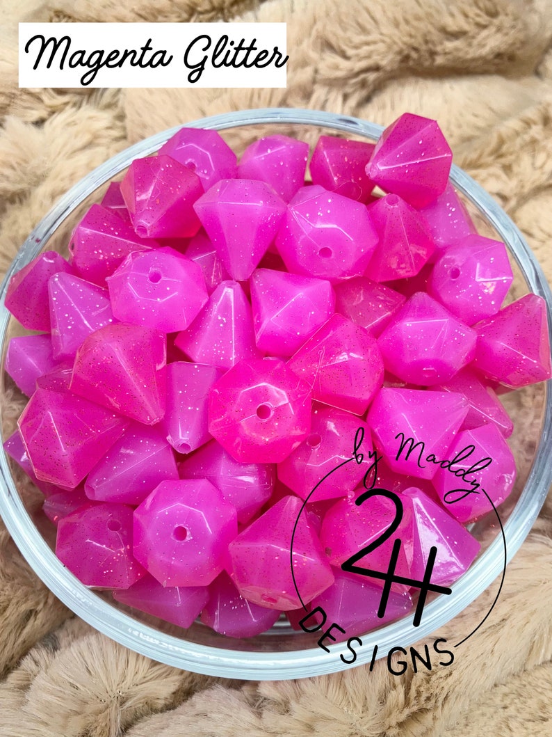 May include: A bowl of magenta glitter diamond-shaped beads. The beads are translucent and have a sparkly finish. The bowl is clear glass and has a white background.