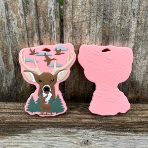 May include: Pink silicone teether with a deer and flying geese design on one side and the text "Baby Bumbles by Molly" on the other side.