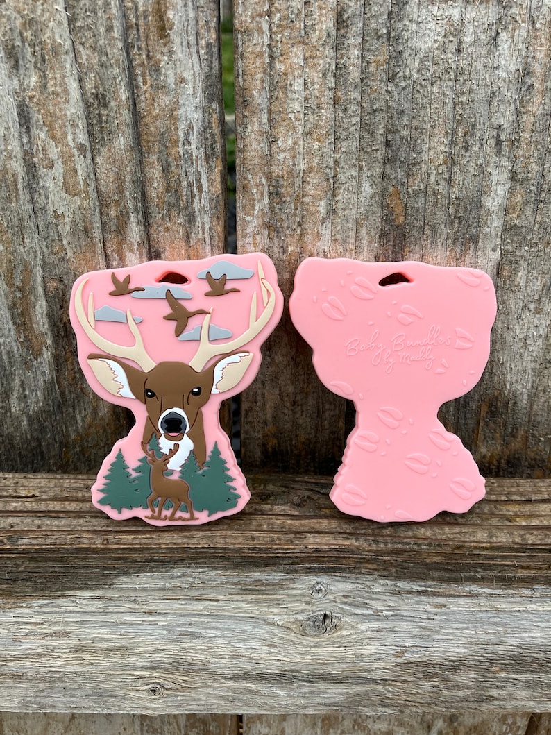 May include: Pink silicone teether with a deer and flying geese design on one side and the text "Baby Bumbles by Molly" on the other side.
