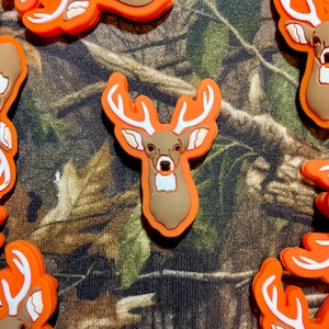 May include: A collection of orange and brown rubber deer head charms with white antlers and eyes. The charms are on a camouflage fabric background.