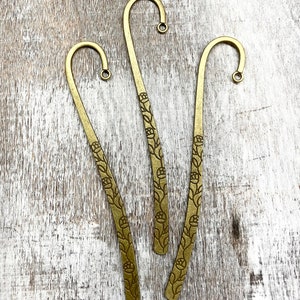 Op de afbeelding: Drie gouden metalen bladwijzers met een bloemenmotief. De bladwijzers zijn haakvormig en hebben een kleine lus aan de bovenkant.