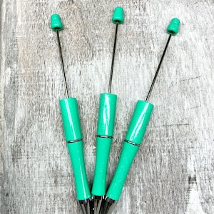 May include: Three teal blue pens with silver accents and a silver tip. The pens are arranged in a triangle shape on a light wood background.