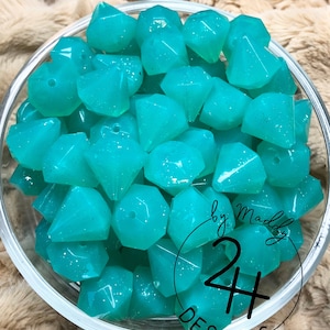 May include: A bowl filled with teal blue, faceted, glitter-filled resin beads. The beads are shaped like diamonds. The text "Ocean Sparkle by Maddy 24 Designs" is visible on the bottom of the bowl.