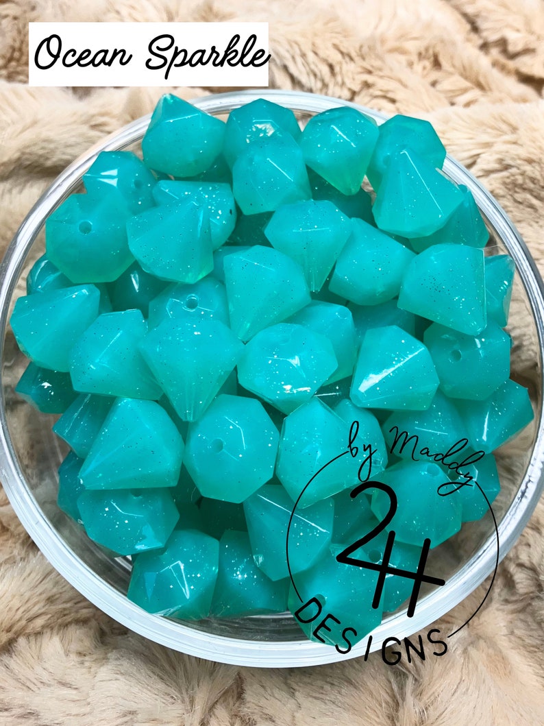 May include: A bowl filled with teal blue, faceted, glitter-filled resin beads. The beads are shaped like diamonds. The text "Ocean Sparkle by Maddy 24 Designs" is visible on the bottom of the bowl.