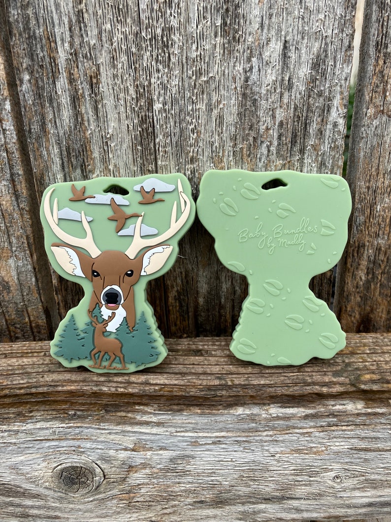 May include: Green silicone teether with a deer and flying geese design. The back side of the teether has a green background with deer footprints. The teether is labeled "Baby Bundles by Maddy".