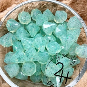 May include: A bowl filled with light green, translucent, faceted beads with a sparkly finish. The beads are shaped like diamonds and have a hole in the center. The text "Jaded Sparkle by Maddy 24 Designs" is visible in the bottom right corner of the image.