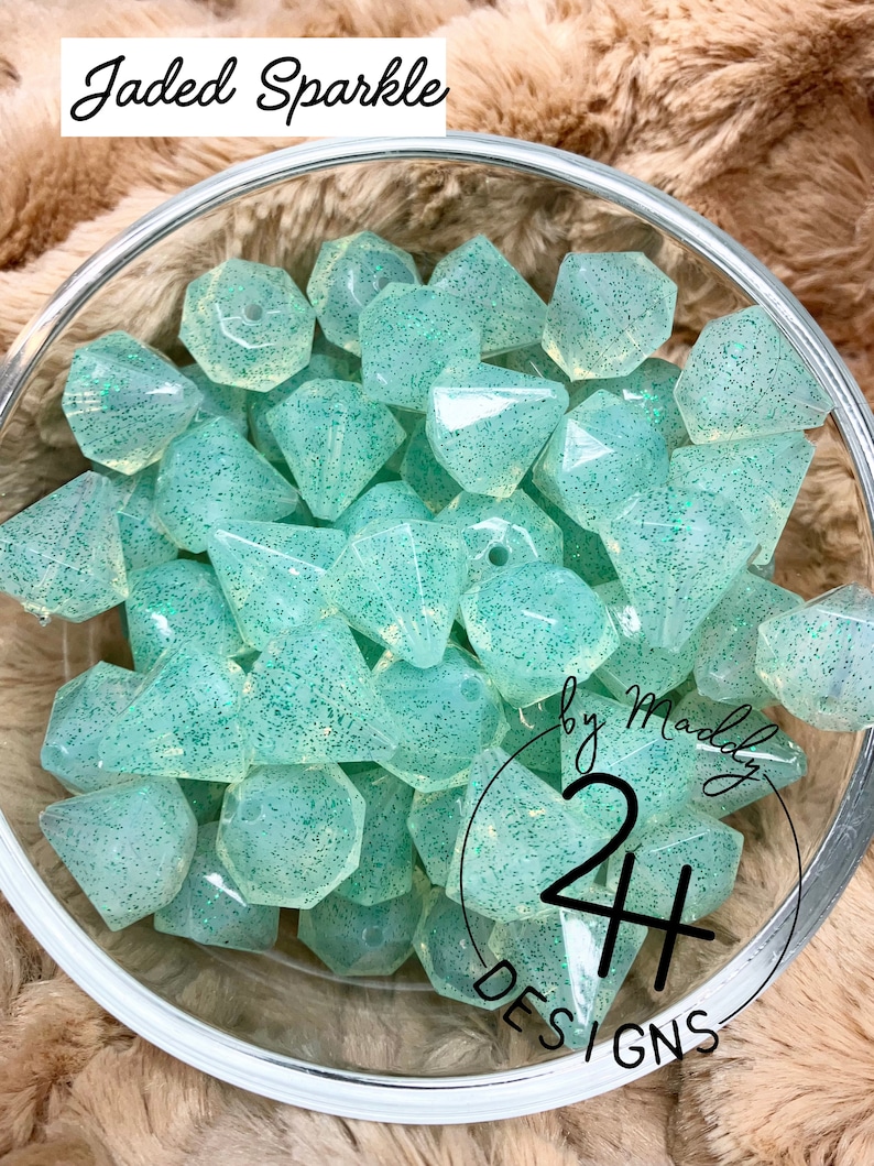 May include: A bowl filled with light green, translucent, faceted beads with a sparkly finish. The beads are shaped like diamonds and have a hole in the center. The text "Jaded Sparkle by Maddy 24 Designs" is visible in the bottom right corner of the image.